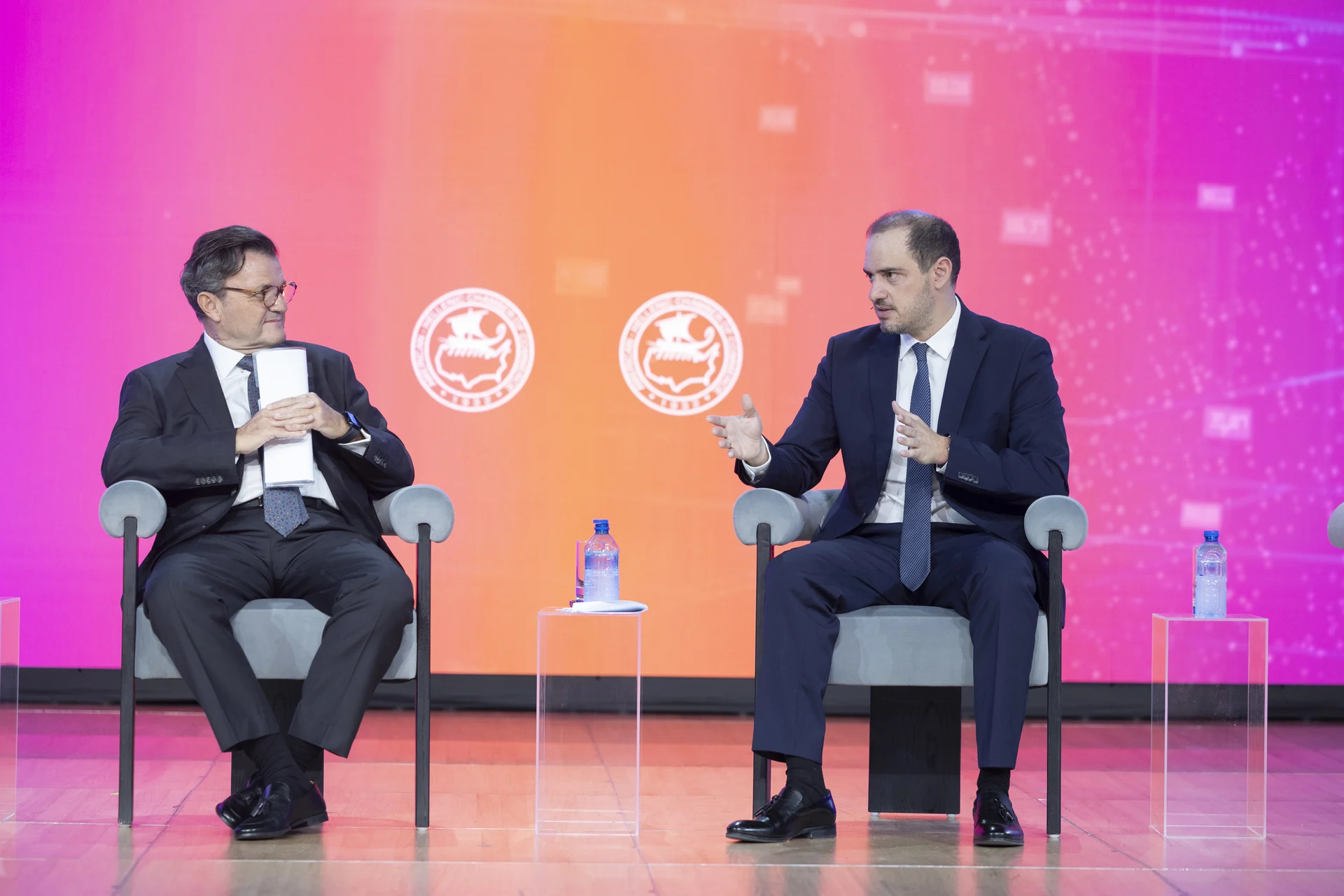 Two speakers seated onstage at the 21st Athens Tax Forum, against a pink and orange conference backdrop.